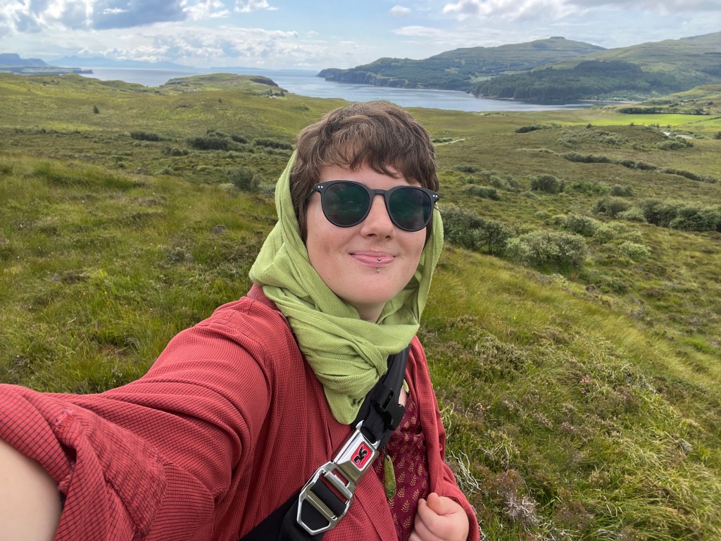 Ruth taking a selfie on Skye in 2025. The background is yellowgreen moorland and the ocean.
